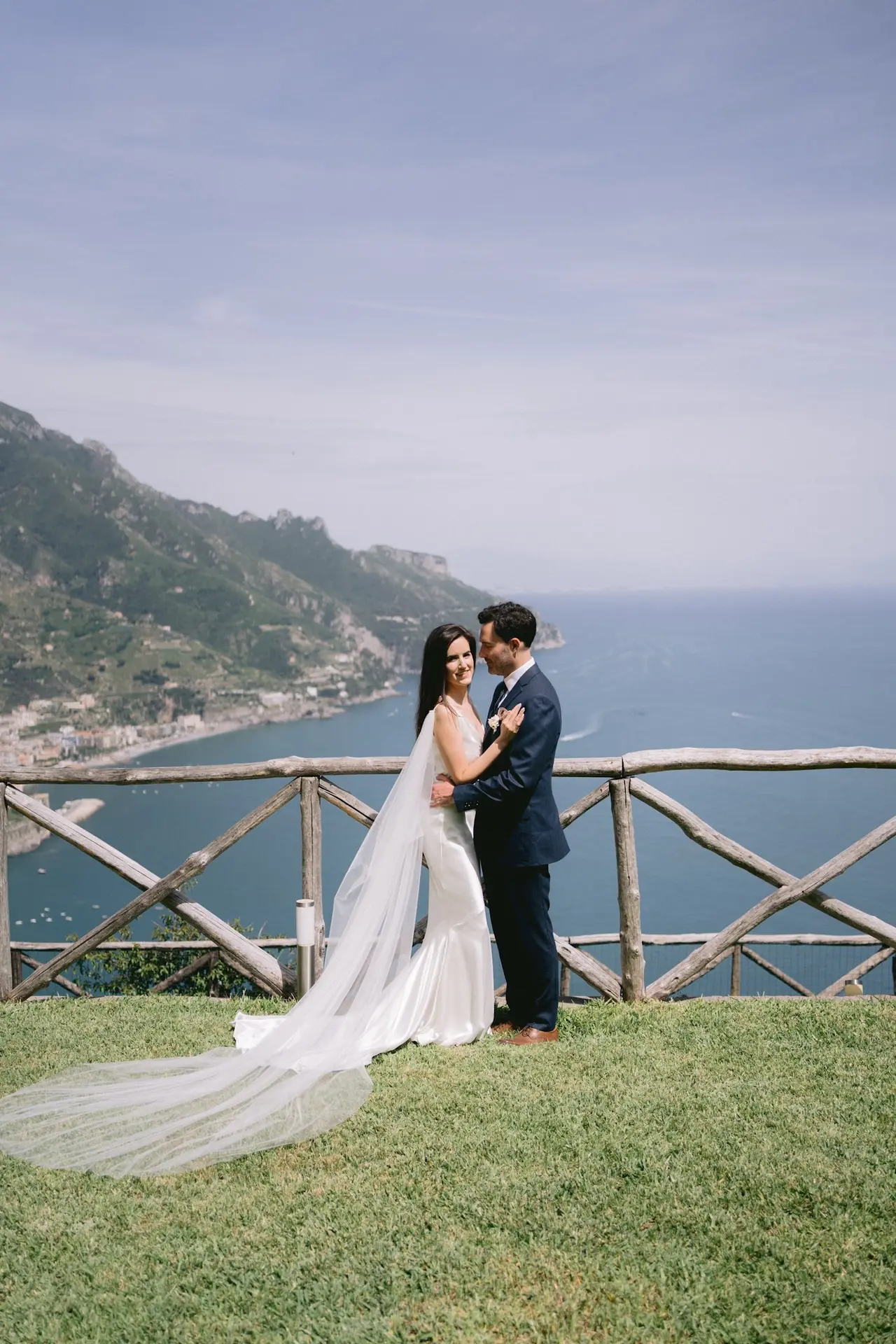 The bride and groom embracing at a coastal viewpoint, her veil trailing across the grass, the coastline behind them