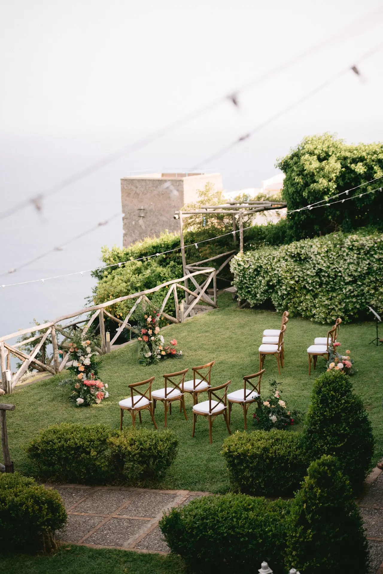 Wider view of the same ceremony seating on the garden lawn, boundary wall, clipped box hedges and string lights strung above