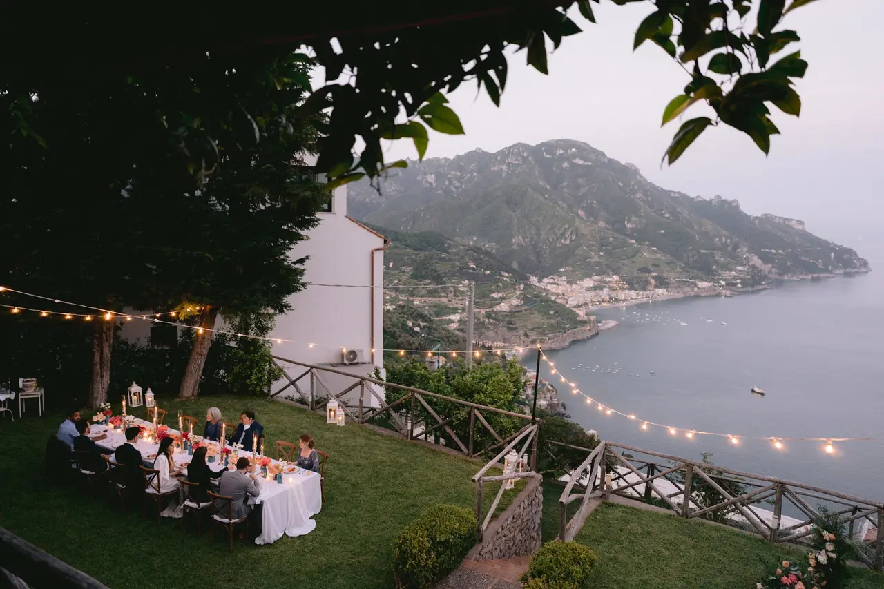 Long dinner table set for a wedding celebration on the Villa Mariella lawn at dusk, string lights overhead and the bay of Maiori below