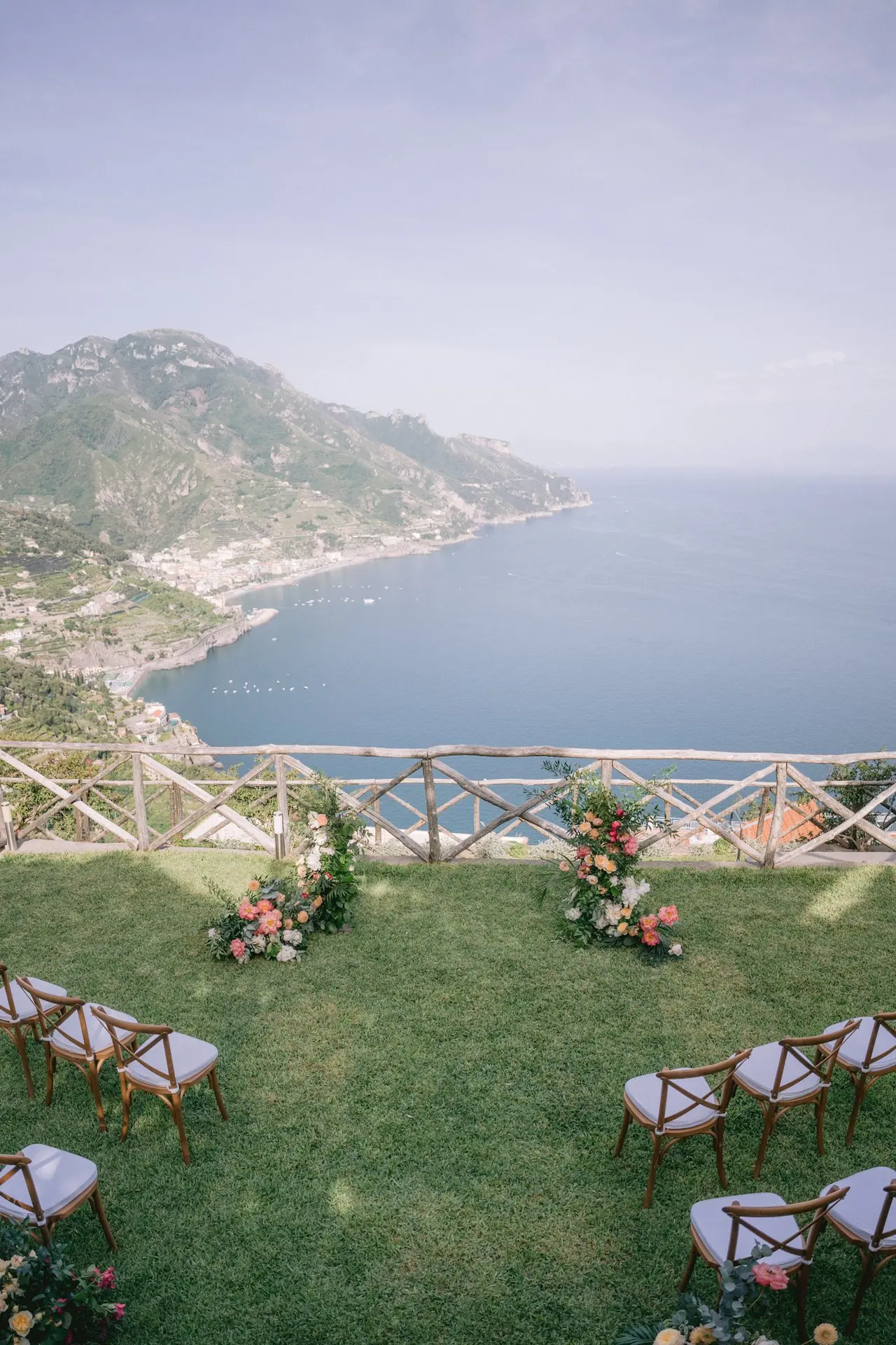 Wooden cross-back chairs set out on the lawn for a ceremony, two coral and white floral arrangements against the chestnut fence, the sea beyond